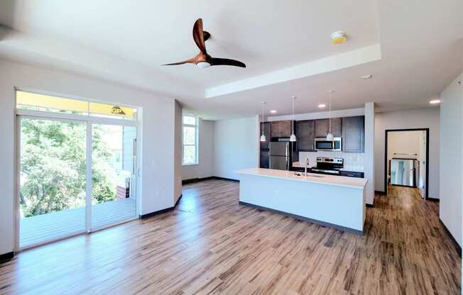 A spacious, empty living room here at Van Buren East with wood-style flooring and modern lighting. A large sliding glass door opens to a private balcony, while the open layout connects to a stylish kitchen area.