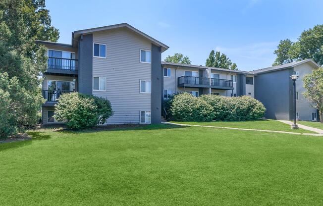 A multi-story residential building with balconies, surrounded by lush green grass and shrubs under a clear blue sky. The structure features a mix of siding colors, with trees in the background, creating a pleasant, inviting atmosphere.