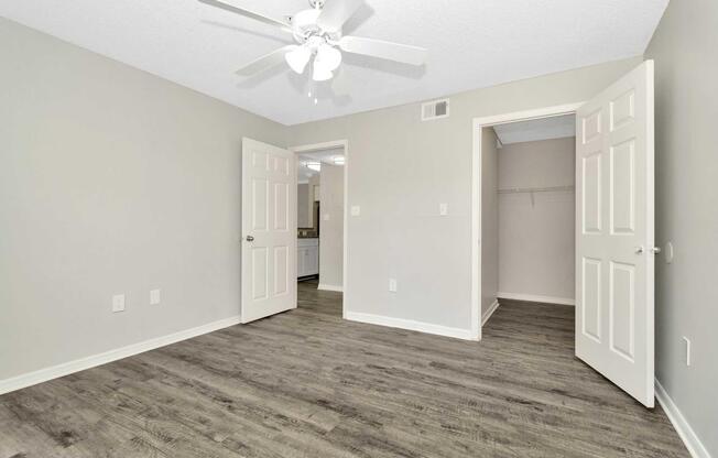 Interior of a room featuring light gray walls and a ceiling fan. There are two open doors leading to adjacent spaces, and the floor is covered with wood-like flooring. The room appears bright and spacious, with a closet visible on one side.