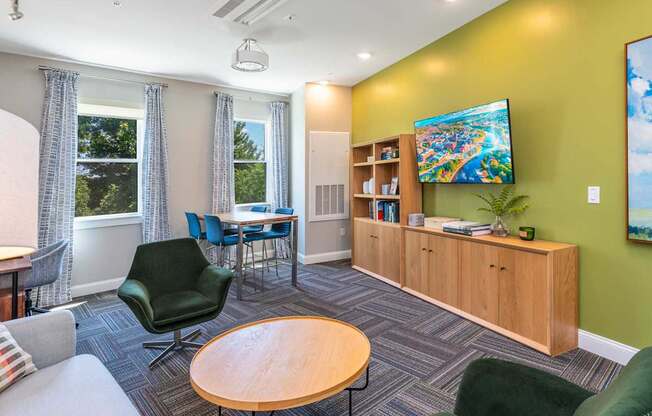 The Residences at Crosspoint cyber lounge in Lowell, MA includes a green accent wall with a mounted flat-screen TV, a wooden storage unit with open shelving, a dark green swivel chair, a round wood coffee table, and a high-top table with blue chairs by two curtained windows overlooking trees.