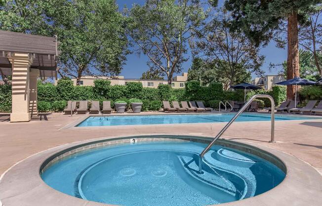 A clear blue swimming pool surrounded by lounge chairs under sunny skies. A hot tub is in the foreground with steps leading in. Lush greenery and trees provide a serene backdrop, along with a shaded area featuring umbrellas. The setting creates a relaxing outdoor atmosphere.