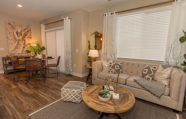 A cozy living room featuring a beige tufted sofa with decorative pillows, a wooden coffee table with plants, and a woven ottoman. Large windows with white blinds allow natural light, and a dining area with a table and chairs is visible in the background. Stylish wall art and indoor plants add a touch of elegance.