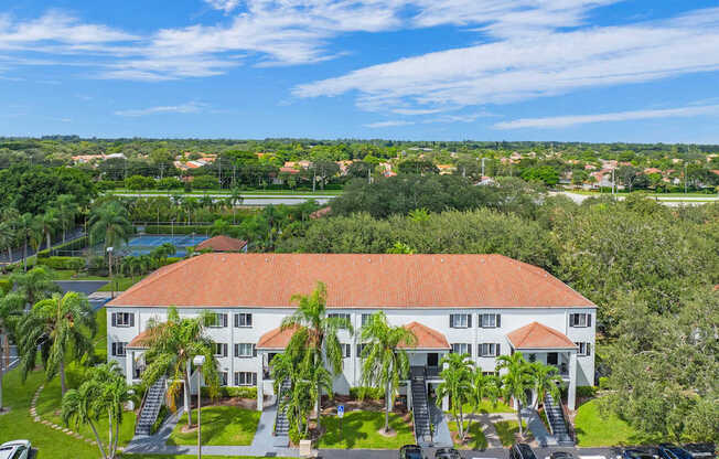 A large white building with a red roof is surrounded by greenery and has a parking lot in front.