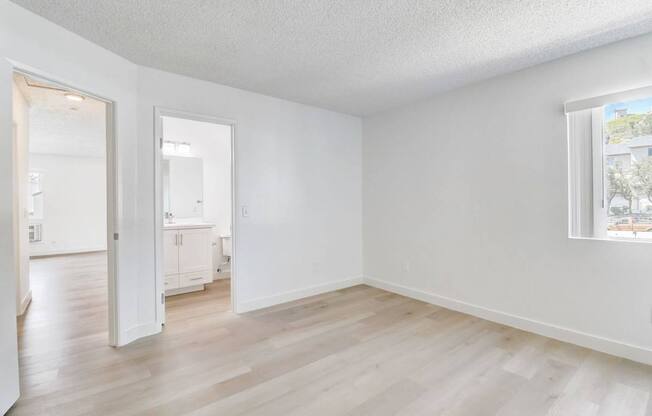 A room with white walls and wooden flooring, with a window showing a building outside.