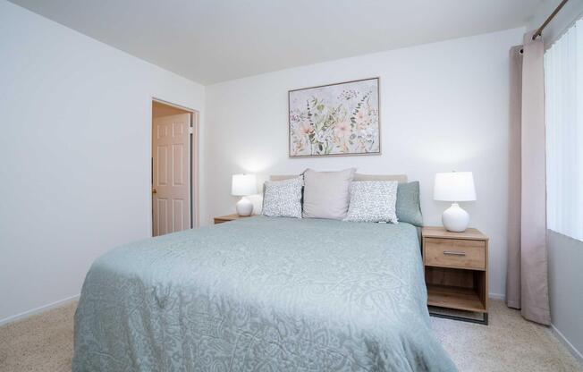 A cozy bedroom featuring a large bed with a light teal covering, two bedside tables with white lamps, and a floral wall art above the bed. The room has neutral walls and carpet, with a window letting in natural light and a door leading to another room.