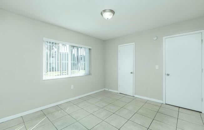 A spacious, empty room with light-colored walls and a tile floor. There's a window with vertical blinds letting in natural light, and two doorsâone on the right and another on the opposite wall. The ceiling features a simple light fixture. The overall atmosphere is bright and minimalist.