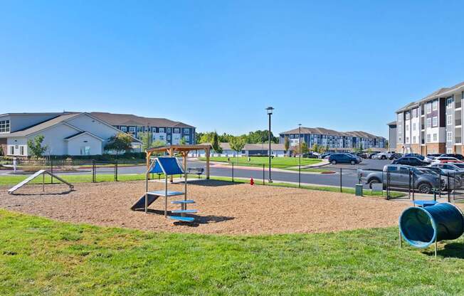 A playground with a slide, swings, and a seesaw in the foreground with a parking lot and buildings in the background.