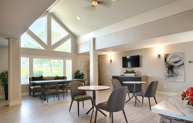 A modern resident gathering space here at Andorra featuring a high vaulted ceiling with triangular windows, round café tables with upholstered chairs, a long dining table with bench seating near large windows, light wood-style flooring, neutral walls with abstract artwork, wall-mounted television, and soft lighting that creates a bright, welcoming atmosphere.