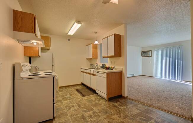 A kitchen with white cabinets and white appliances with a living room with a door to a balcony. Fargo, ND Eagle Run Apartments.
