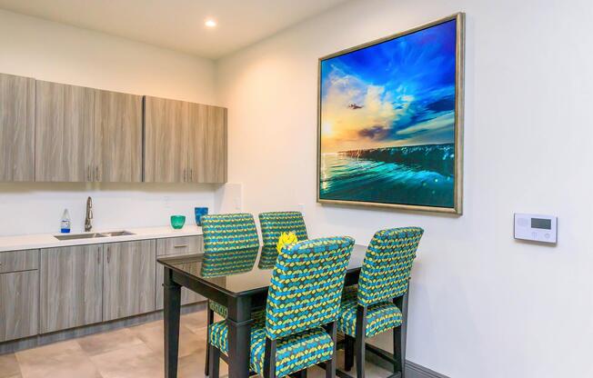 A modern kitchen featuring light wood cabinets and a minimalist design. There is a black dining table with four colorful upholstered chairs displaying a vibrant pattern. A large framed artwork depicting a scenic sunset over the ocean is hung on the wall, adding a touch of color and warmth to the space.