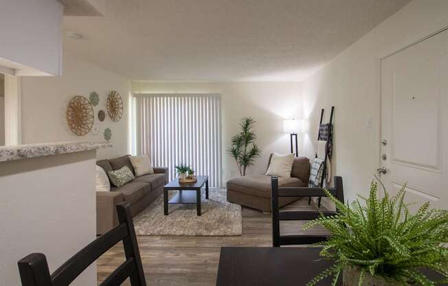 View of the living room with a couch, chaise lounge and a potted plant from the dining room of a 515 square foot 1 bedroom, 1 bath apartment home at Canyon Creek Apartments in Dallas Midtown neighborhood of Dallas, TX.