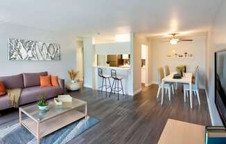 A modern living room with a grey sofa, a glass coffee table, and a dining area in the background.