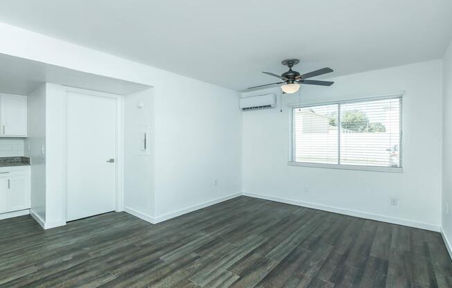 A bright, airy room with white walls and a ceiling fan. The wood-patterned floor adds warmth. To the left, there is a doorway leading to another space, and a window on the right allows natural light to fill the room, framed by white blinds. A small air conditioning unit is also visible.