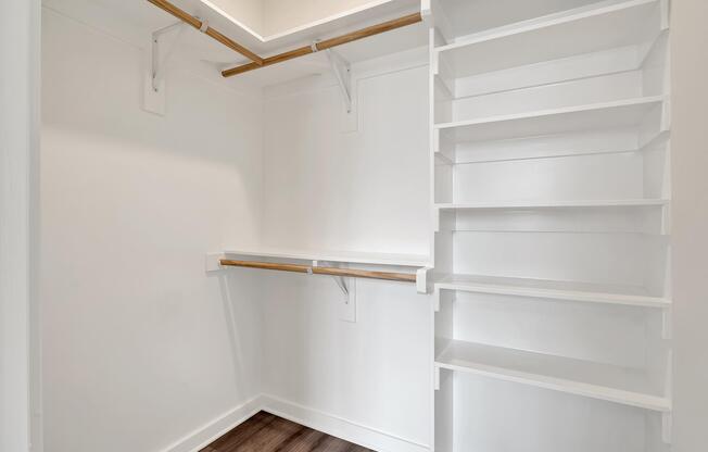 Empty walk-in closet featuring white walls, wooden shelving, and a spacious layout with two hanging rods for clothes and several open shelves for storage. The flooring is a dark wood, providing a contrast to the bright, uncluttered design.