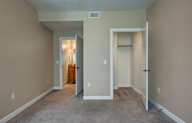 A bedroom here at Bridges with beige walls, gray carpet, an open white closet, and a doorway leading to a bright bathroom vanity area with warm wood cabinetry and mirror lighting.