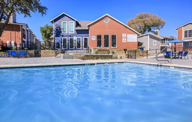 A calm swimming pool in the foreground reflecting the clear blue sky, with a modern, multi-colored building in the background. The building features large windows and outdoor seating areas. Surrounding the pool are lounge chairs and tables, creating a relaxing outdoor space.