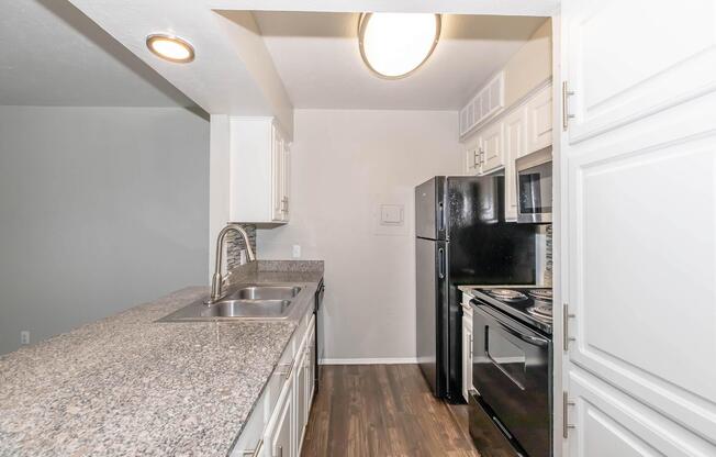 Bright and modern kitchen featuring granite countertops, white cabinets, a stainless steel sink, and black appliances including a refrigerator and oven. The open layout is well-lit with overhead lighting, and the flooring is a warm wood tone, creating an inviting atmosphere.