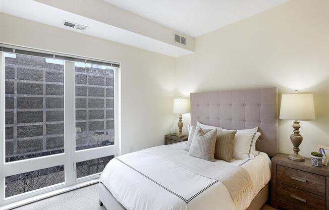 A bedroom with a large bed, a nightstand with a lamp, and a window with a view of a cityscape.