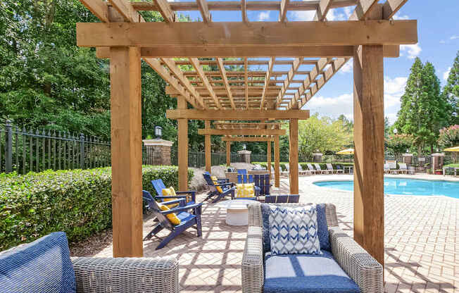 A wooden pergola over a poolside seating area with blue cushioned chairs.