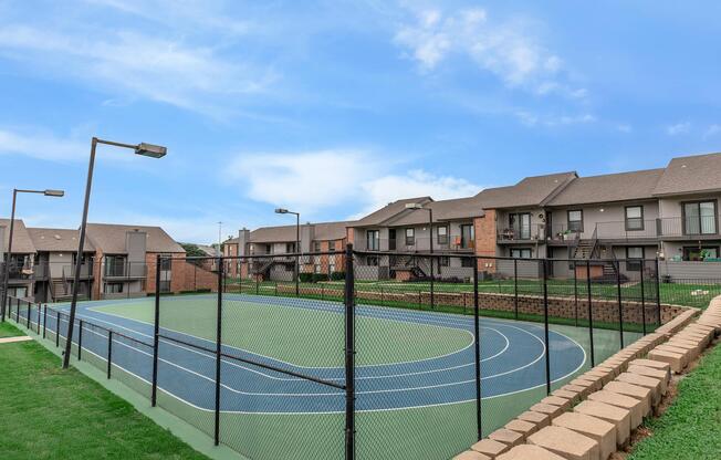 A view of an outdoor tennis court surrounded by a fence, with several residential buildings in the background. The court features green and blue surfaces, and the area is well-maintained with grass and lighting fixtures. The sky is clear and blue, creating a pleasant atmosphere.