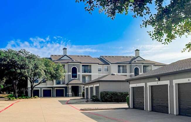 Exterior view of Saxony at Chase Oaks Apartments in Dallas, TX, showcasing three-story buildings with arched entryways, private balconies, and attached garages, surrounded by mature trees and a spacious driveway.