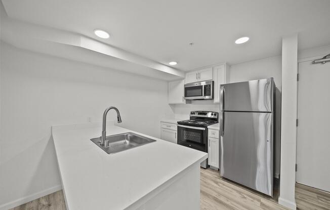 Modern kitchen with white cabinetry, stainless steel appliances including a microwave, oven, and refrigerator. A large island with a sink is in the foreground, and the space features light wood flooring and recessed lighting for a bright, open feel.