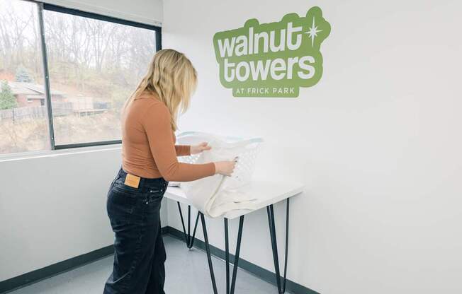 A woman is setting up a table in a room with a sign that says "walnut towers at Frick Park".