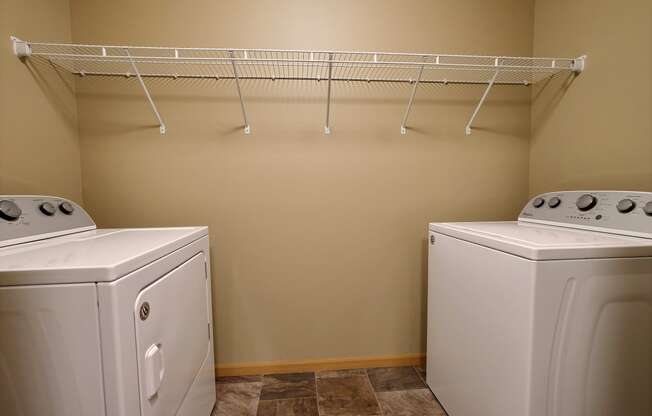 two washers and a dryer in a laundry room with a rack on top. Fargo, ND Urban Plains Apartments