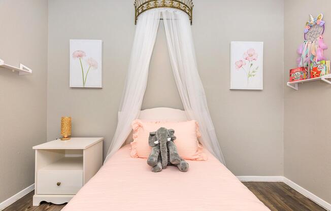 A cozy bedroom featuring a single bed with a light pink duvet, adorned with a gray stuffed elephant. Above the bed, two floral art prints hang on the wall. A white nightstand sits beside the bed, and delicate shelves display decorative items. A sheer white canopy adds a whimsical touch to the space.