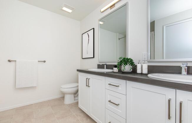 Modern bathroom featuring a double vanity with two sinks, a large mirror, and contemporary fixtures. The room has light-colored walls, a towel rack with a white towel, a small plant arrangement, and a toilet in the corner. The neutral tile floor adds to the clean and spacious feel of the space.