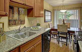 A bright kitchen here at Whispering Cedars with light brown cabinets, granite countertops, and a stainless steel sink, featuring black appliances and an adjacent dining area with a wooden table, cushioned chairs, and sliding glass doors bringing in natural light.