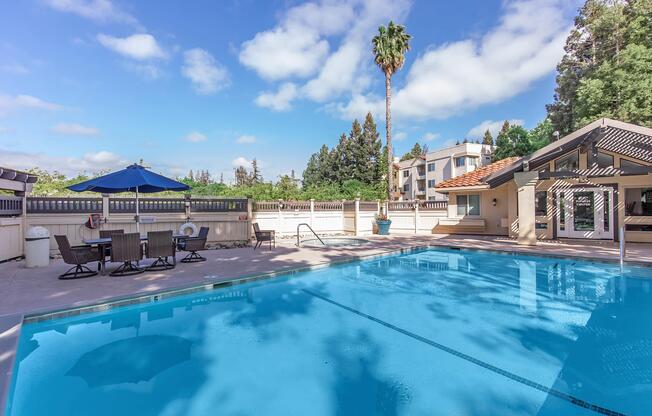 A serene outdoor swimming pool surrounded by a patio area with lounge chairs and a dining set under an umbrella. Lush greenery and palm trees frame the scene, while a clear blue sky adds to the inviting atmosphere. The modern building in the background completes the tranquil setting.