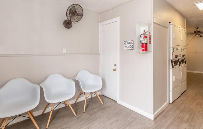 A minimalist waiting area featuring three white chairs with wooden legs, a wall-mounted fire extinguisher, a "No Smoking" sign, and a laundry area with multiple machines. The walls are painted a light color, with a ceiling fan visible in the background.