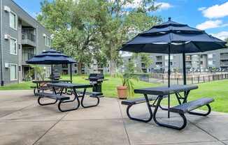 A black and white picnic table with a blue umbrella is in the foreground of a courtyard with apartment buildings in the background.