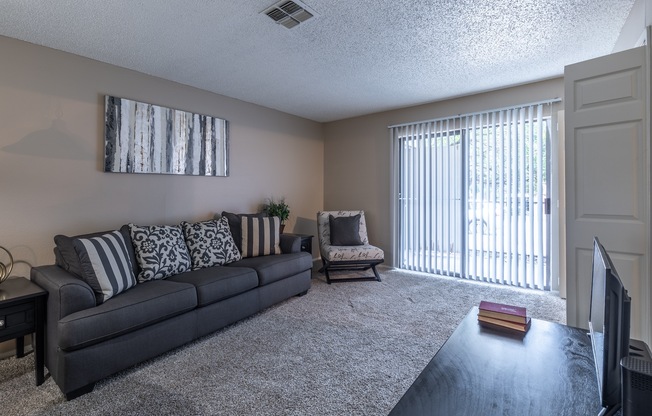A living room with a grey couch, a small table, a television, and a window with blinds.