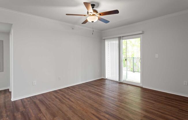 A spacious, empty living room with light gray walls and hardwood flooring. It features a ceiling fan and large sliding glass doors that lead to a balcony. Natural light floods the room, highlighting the simplicity and openness of the space.