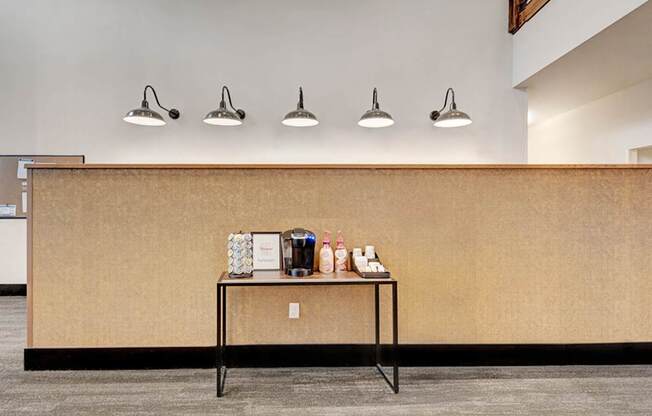 A welcoming refreshment station here at The Park featuring a coffee maker, organized condiments, and modern wall sconces above a warm accent wall, creating a convenient and inviting community amenity.