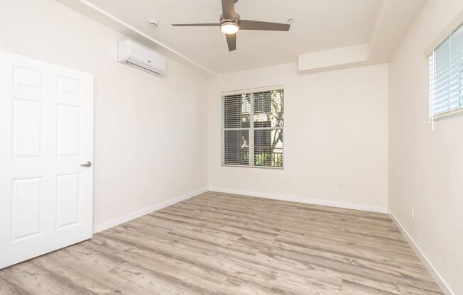 A spacious, empty room with light-colored walls and a wooden floor. It features a ceiling fan, a window with blinds, and a white door. Natural light illuminates the space, highlighting its clean and modern design.
