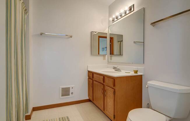 A clean bathroom here at Arbor Ridge featuring a light wood vanity, large mirror, and bright lighting. Neutral flooring and striped shower curtain create a fresh, welcoming feel.