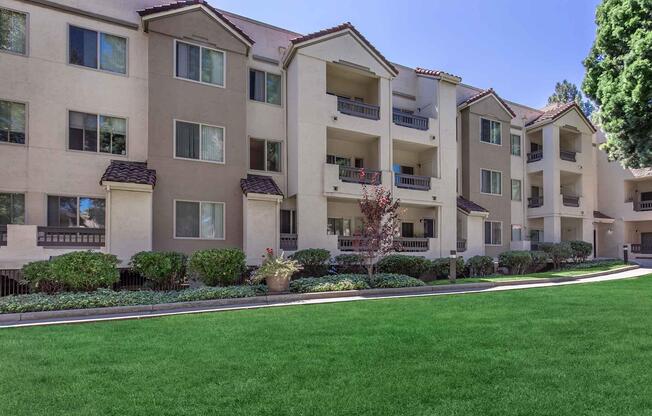A well-maintained apartment community featuring multiple beige buildings with balconies, surrounded by manicured lawns and shrubs. The scene is set on a bright, sunny day, highlighting the greenery and inviting atmosphere of the outdoor space.