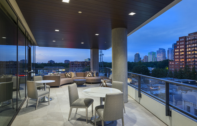 An open-air loggia with a view of the city skyline at dusk, a wraparound glass railing, a seating area with a curved couch and armchairs, and multiple sets of white tables and chairs.