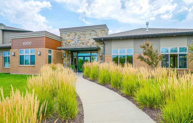 A sidewalk surrounded by tall grass leasing to a modern stone and brick clubhouse with large windows.