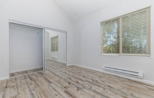 Empty room featuring light-colored walls, a large mirror closet with sliding doors, a window with blinds allowing natural light, and a wood-style floor. The space is bright and airy, suitable for various design possibilities.