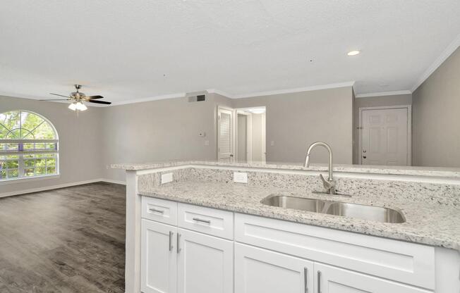 Modern kitchen with granite countertops and a sink, opening to a spacious living area. Natural light enters through a large arched window, highlighting the neutral-colored walls and new flooring. The room features a ceiling fan and leads to a hallway with a doorway visible in the background.