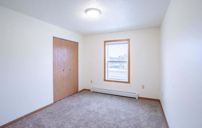 A room with a carpeted floor, a door, a window, and a small radiator. Fargo, ND Park Place Apartments