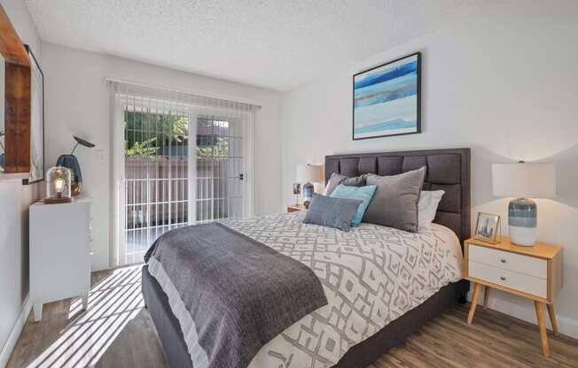 One Bedroom Apartments in Hayward CA - Glen Oaks - Spacious Bedroom with Wood-Style Flooring, a Bed, Wooden Nightstands with Lamps, White Dresser, and Large Window/Sliding Door with Patio Access