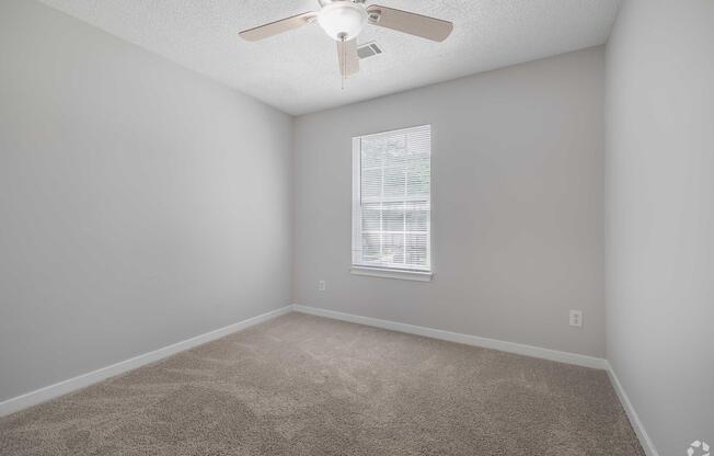 A vacant room with light gray walls and carpeted flooring. There's a ceiling fan with light fixtures and a window allowing natural light to enter. The space appears clean and uncluttered, ideal for personal customization or furniture arrangement.