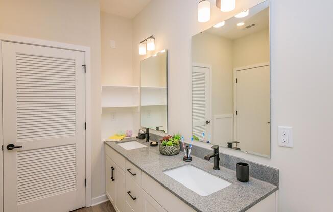 A modern bathroom featuring a double sink vanity with a grey countertop, two mirrors above the sinks, and a contemporary light fixture. There are decorative elements like a potted plant and neatly arranged toiletries. The walls are painted in a light color, and a door with horizontal slats leads to another space.