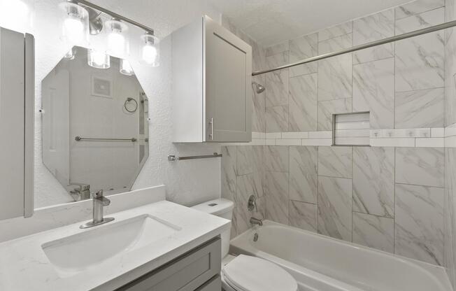 A modern bathroom featuring a white bathtub, gray cabinetry, a marble-patterned tile wall, and a large mirror above the sink. The lighting consists of four glass bulb fixtures, and there are chrome fixtures and a towel rack visible. The overall design is clean and contemporary.