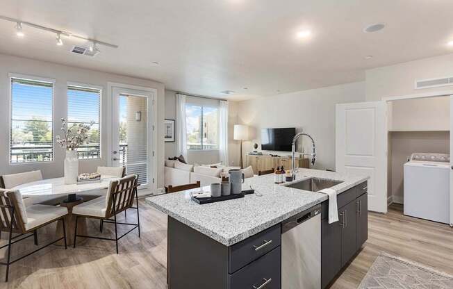 A bright open living space here at Reflect at Dobson Ranch featuring a modern kitchen with dark cabinetry, a large granite-style island with an undermount sink and bar seating, and wood-style flooring, flowing into a dining area and inviting living room with a neutral sectional, natural light, and access to a private balcony.